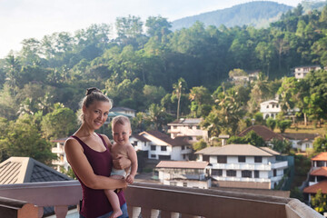 Portrait of mom with baby on balkony in tropical country at mountain background. Woman with child in summer travel vacation. Concept of motherhood and family recreation with children. Copy text space