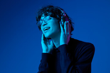 Wall Mural - Teenage man wearing headphones listening to music and dancing and singing with glasses, hipster lifestyle, portrait blue background, neon light, style and trends, mixed light, copy space