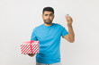 Portrait of puzzled attractive man wearing blue T- shirt standing choosing between two present boxes, choosing little gift box with disappointment. Indoor studio shot isolated on gray background.