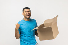 Portrait Of Extremely Happy Unshaven Man Wearing Blue T- Shirt Standing With Cardboard Box In Hand, Clenches Fist, Celebrating Success. Indoor Studio Shot Isolated On Gray Background.