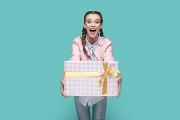 Poster - Portrait of excited amazed teenager girl with braids wearing pink jacket giving wrapped present box with gold ribbon, congratulating. Indoor studio shot isolated on green background.