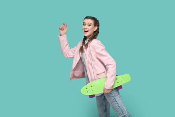 Wall Mural - Portrait of extremely happy funny teenager girl with braids wearing pink jacket standing with raised arm, holding skateboard in hands. Indoor studio shot isolated on green background.