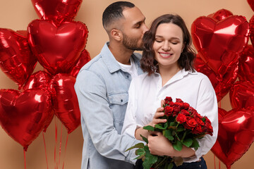 Poster - Happy couple celebrating Valentine's day. Beloved woman with bouquet of red roses near heart shaped air balloons on beige background