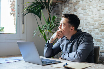 Wall Mural - Thoughtful businessman touching chin, pondering ideas or strategy, sitting at wooden work desk with laptop, freelancer working on online project, student preparing for exam at home
