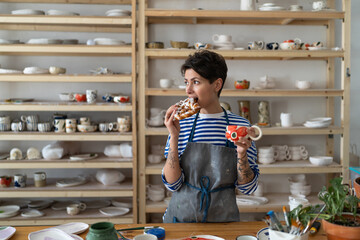 Wall Mural - Relaxed woman in apron drink coffee from handmade ceramic cup and has snack in pottery studio. Tattooed female artist has break at workplace after work. Small business and art concept