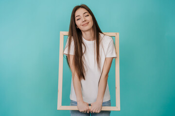 Cute cheerful young woman in white t-shirt and jeans holds frame for Canvas smiling wide stands over turquoise studio backdrop with empty space. Attractive Italian girl got into wooden frame. Mockup