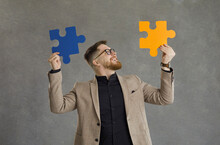 Happy Creative Guy In Suit And Modern Hipster Glasses Joins Together Orange And Blue Jigsaw Puzzle Pieces Standing Isolated On Grey Background. Concept Of Finding Good Professional Business Solution