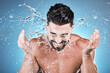 Water splash, hands and a man model washing his face in studio on a blue background for hygiene or hydration. Bathroom, skincare and cleaning with a handsome young male splashing his skin for beauty