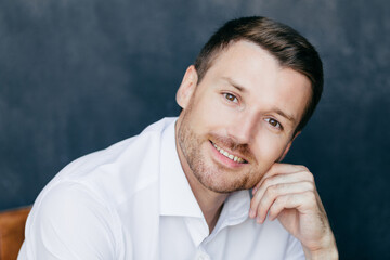 Wall Mural - Photo of young male entrepreneur with bristle, keeps hand on cheek, dressed in elegant white shirt, poses against dark background, has toothy smile. Elegant handsome young successful man indoor