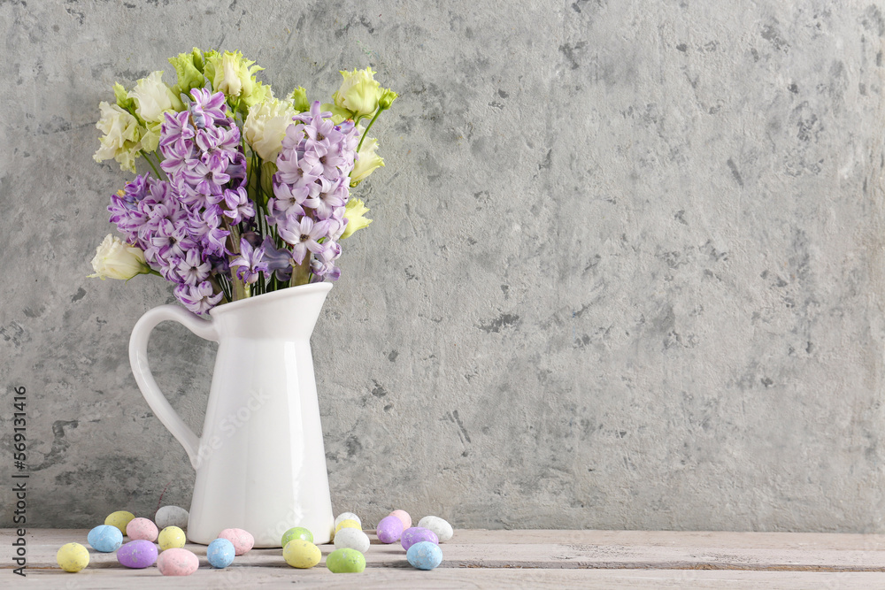 Jug with beautiful flowers and Easter eggs on wooden table near light wall