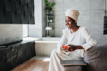 Happy African woman in white dress and turban holds phone sitting at home on couch. Happy African American businesswoman laughs looks away being satisfied. People candid emotions. Woman in hotel.