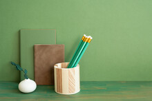 Green And Brown Diary Notebook, Pencil Holder, Plant On Wooden Desk. Green Wall Background. Workspace, Copy Space