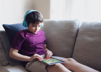 Wall Mural - Education concept,High key portrait primary school Kid reading E-book on tablet for homework,Child wearing headphone playing game online on internet,boy sitting on sofa watching cartoon on digital pad