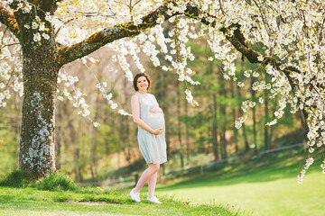 Outdoor maternity portrait of happy young pregnant woman standing next to blooming tree, springtime