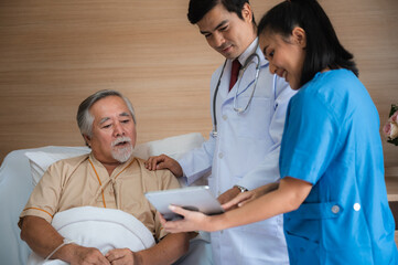 Smiling senior male patient lying and sitting on bed resting while nurse and doctor in lab coat and stethoscope showing digital tablet screen to man in a hospital room