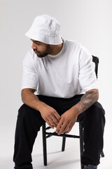 Wall Mural - Attractive african american man with a beard dressed in a white t-shirt and panama sits on a chair.