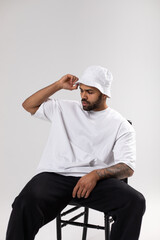 Wall Mural - Attractive african american man with a beard dressed in a white t-shirt and panama sits on a chair.