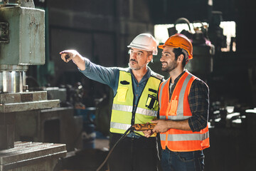 Poster - Senior male engineer training and explaining work to new employee wearing vest and safety jacket with hardhat helmet while pointing towards machine in factory and giving instructions