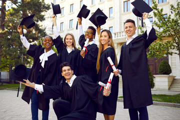 Wall Mural - Young multinational people university students dressed in black gown raise their hats rejoicing in successful completion higher education with excellent grades standing in front of college building