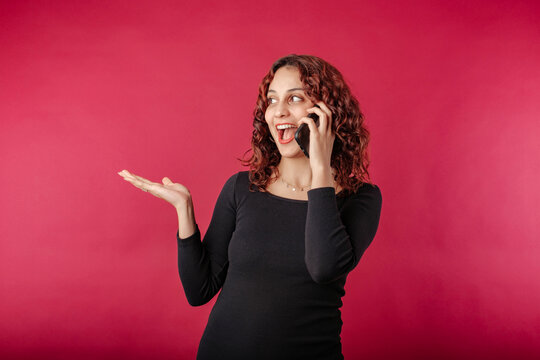 Young redhead girl smiling happy wearing black ribbed dress isolated over red background on the phone holding on palm outstretched while looking at the empty copy space.