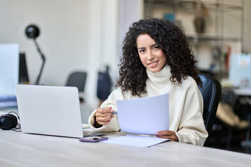 Young smiling latin business woman manager accounting analyst checking bills, analyzing sales management report, taxes financial data documents or marketing papers working in office using laptop.