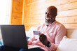 © Wavebreak Media - Bald african american senior man with credit card doing online shopping over laptop in log cabin