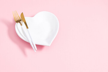 Valentine's Day concept. White empty heart shaped plate and cutlery fork and knife on isolated pastel pink background. Flat lay, top view, copy space