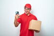 © Reza - Asian courier man in red uniform smiling look at his smartphone while holding cardboard box parcel