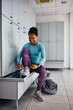 © Drazen - Happy black female athlete tying shoelace on sneakers in locker room at gym.