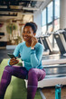 © Drazen - Happy African American sportswoman using earbuds and cell phone in gym while looking at camera.