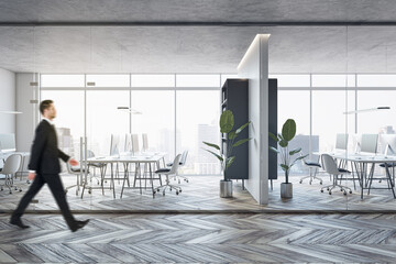 Poster - Side view of young businessman walking in modern glass office interior with wooden flooring, furniture, window with city view and other objects.