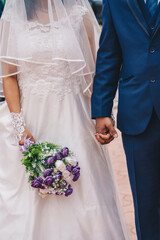 Cropped photo of bride and groom holding hands on wedding day. The bride wearing a beautiful white lacy gown with veil, roses in hand and the bride groom wearing blue suit 