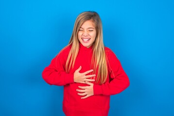 Wall Mural - caucasian teen girl wearing red sweater over blue studio background keeps hands crossed, laughs out loud at good joke, wears casual clothes.