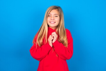 Wall Mural - Dreamy charming caucasian teen girl wearing red sweater over blue studio background with pleasant expression, keeps hands crossed near face, excited about something pleasant.