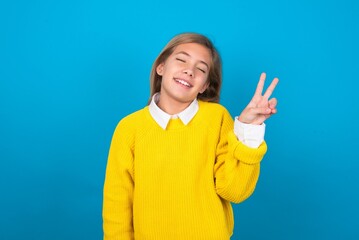 caucasian teen girl wearing yellow sweater over blue studio background smiling with happy face winking at the camera doing victory sign. Number two.