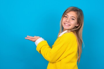 Wall Mural - caucasian teen girl wearing yellow sweater over blue studio background pointing aside with hands open palms showing copy space, presenting advertisement smiling excited happy