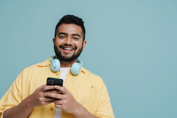 Wall Mural - Smiling indian guy using mobile phone while standing isolated over blue wall