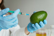 Young woman wearing scientist uniform injecting on avocado during experiment with vegetables in laboratory.