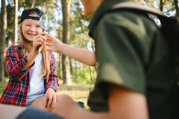 Wall Mural - Group of curious happy school kids in casual clothes with backpacks exploring nature and forest together on sunny autumn day, girl holding magnifying glass and looking at fir cone in boy hands.