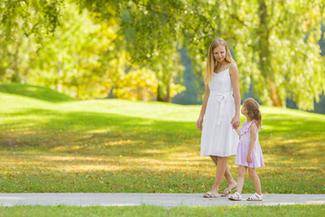 Wall Mural - Little daughter and young adult mother in dresses speaking and walking on sidewalk at city park. Spending time together in beautiful warm sunny summer day. Side view.