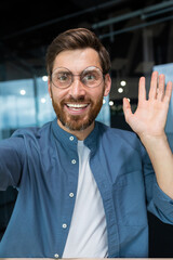 Mature man in office wearing shirt and glasses smiling and looking at smartphone camera, businessman taking selfie and talking on video call using smartphone, programmer waving hand greeting gesture.