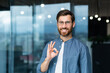 © Liubomir - Portrait of a successful mature businessman inside the office, a man with a beard and glasses in a blue shirt is smiling and looking at the camera, showing the ok gesture with his hand.