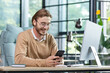 © Liubomir - A successful young businessman, programmer, freelancer sits in a modern office at a table, uses the phone, plays online games, texts, reads messages, smiles.