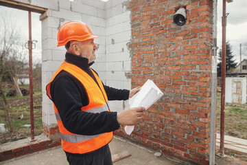 Senior man engineer or construction worker in hardhat looking at blueprints at building new modern house. Male architect or contractor checking plans at construction site. Copy space