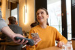 © Folenial - Portrait of a female client smiling and using smartphone to pay for coffee to crop waiter while sitting at table in the morning in a light cafe indoors. Lifestyle concept