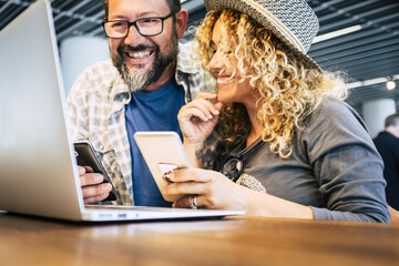 Wall Mural - Modern happy youth couple working together with smartphone and laptop. Portrait of cheerful man and woman using computer and phones at the desk in a cafe or coworking space. Online business people