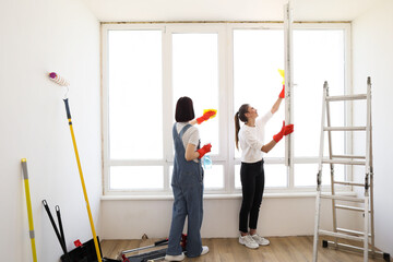 Beautiful young women in rubber gloves with detergent and rag washing windows together. Attractive caucasian girls making house cleaning after repair.