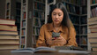 © Yuliia - Young serious asian woman student sitting in library at desk searching information on Internet using mobile phone preparing for exam use free wifi on smartphone take break in learning reading email