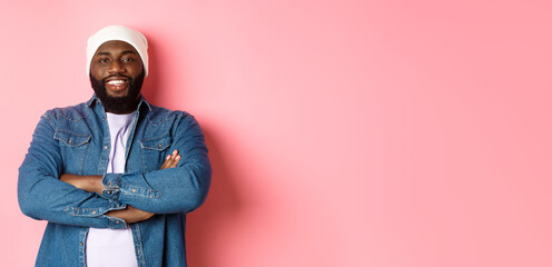 Wall Mural - Handsome hip hop style Black man in beanie and denim shirt, smiling confident, cross arms on chest and staring at camera on pink background