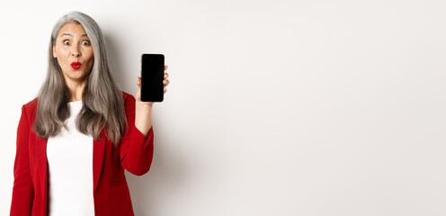 Wall Mural - Amused senior woman in red blazer, looking fascinated and showing blank smartphone screen, standing over white background